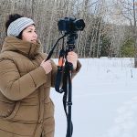 Photo of Lia Taranchansky standing outside in the snow with a brown jacket on, holding a tripod and a camera. 
